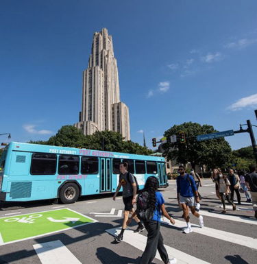 People using a crosswalk in front of a bus, with Pitt's Cathedral of Learning in the background.